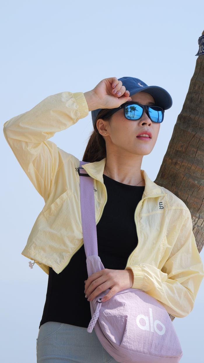 Young woman wearing sunglasses and cap standing by a palm tree under clear blue sky.